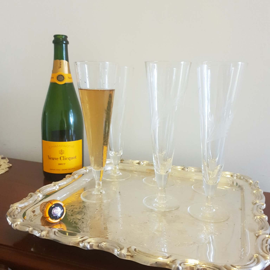 A green Veuve Clicquot champagne bottle with a yellow label stands beside a large ornate silver tray holding six clear champagne flutes, one partially filled with golden champagne, against a plain light wall.
