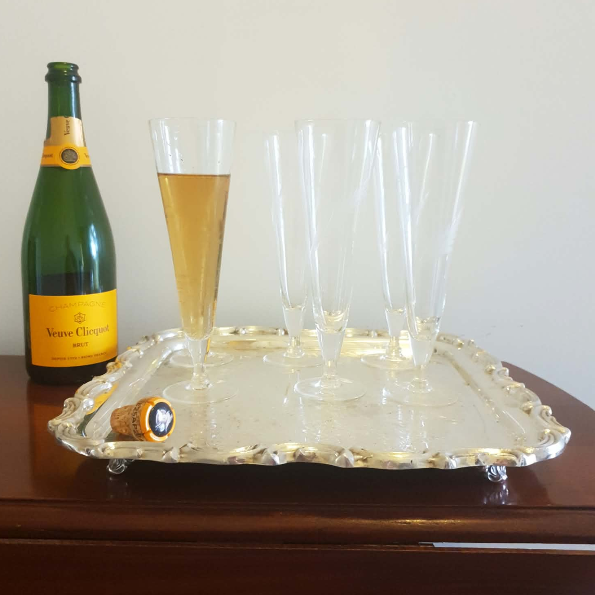 A green Veuve Clicquot champagne bottle with a yellow label stands beside a large ornate silver tray holding six clear champagne flutes, one partially filled with golden champagne, against a plain light wall.