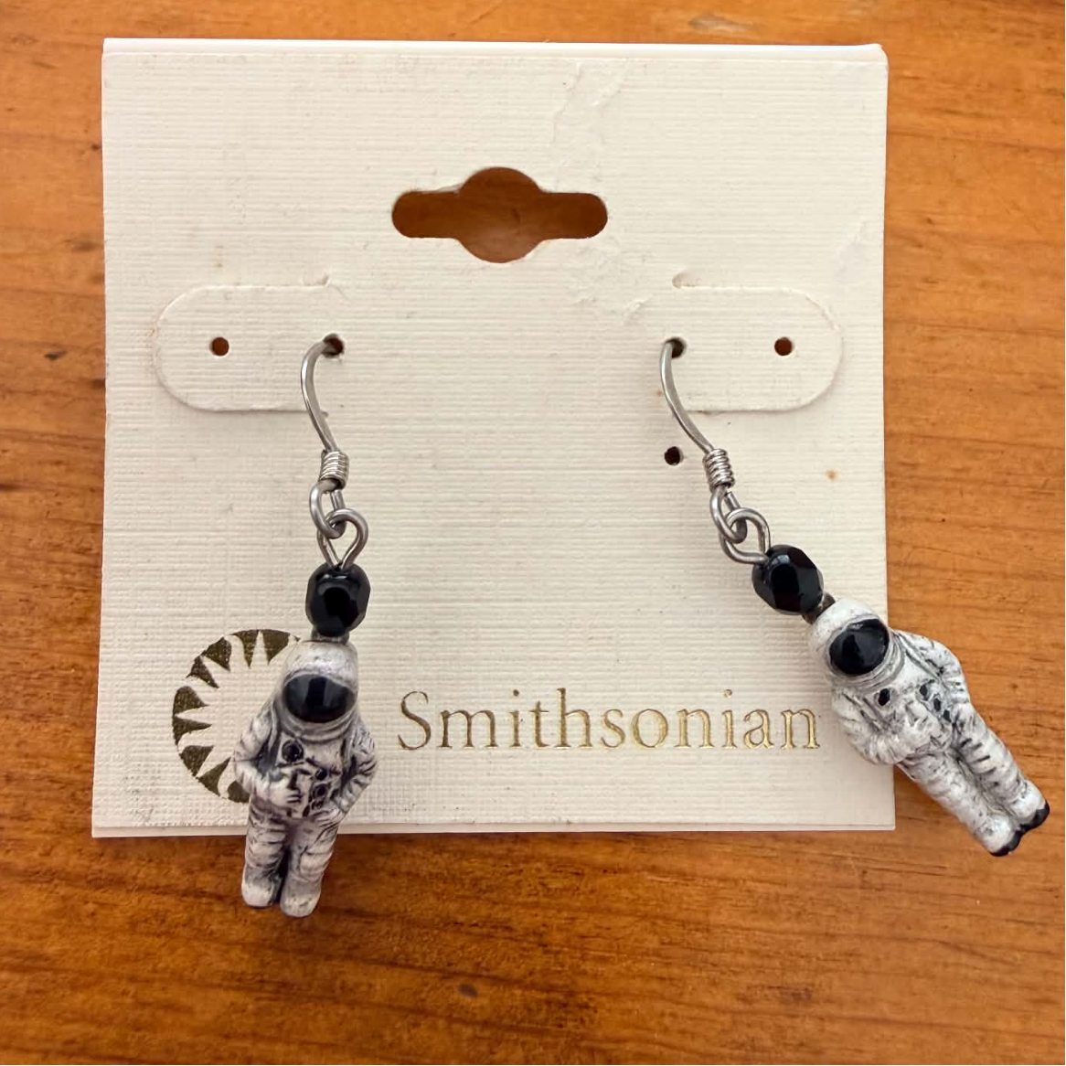 A pair of astronaut dangling earrings displayed on a cream-colored Smithsonian card, set against a warm wooden background.